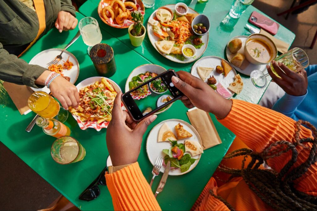 Uma mesa de restaurante com pessoas comendo e uma tirando foto dos alimentos com um celular.
